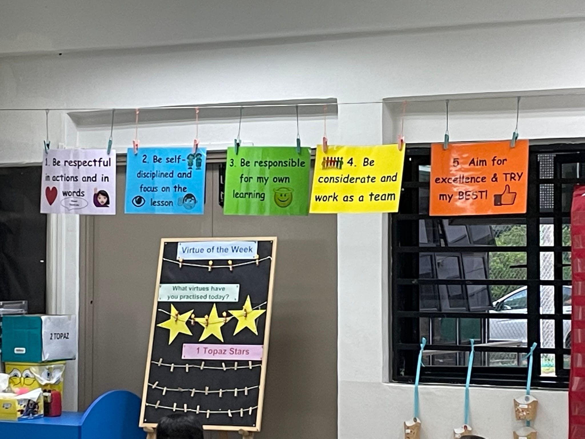 Five colorful classroom rule cards hang clipped to a rail, listing rules like be respectful and aim for excellence.